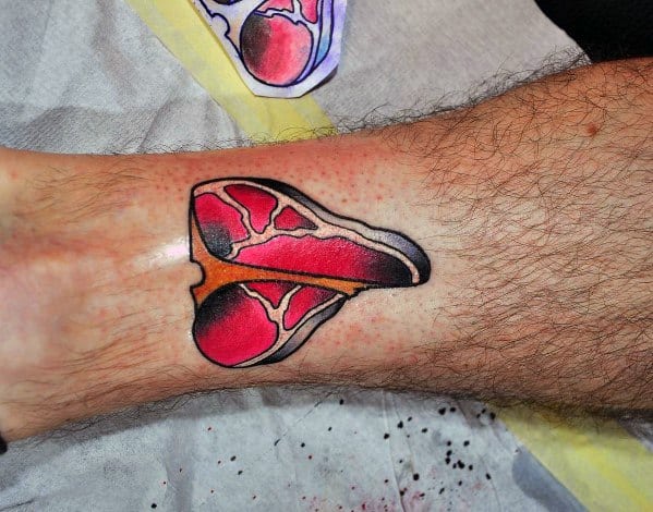 Tattoo of a stylized T-bone steak with bold red and black shading, placed on the ankle for a striking and unique meat-inspired design