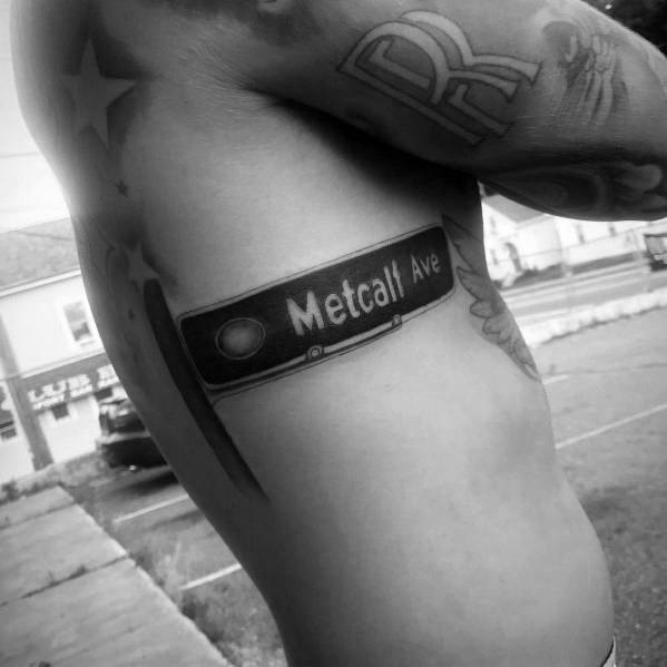 Bold black and grey tattoo of a 'Metcalf Ave' street sign on the ribcage, designed with a realistic, high-contrast style.