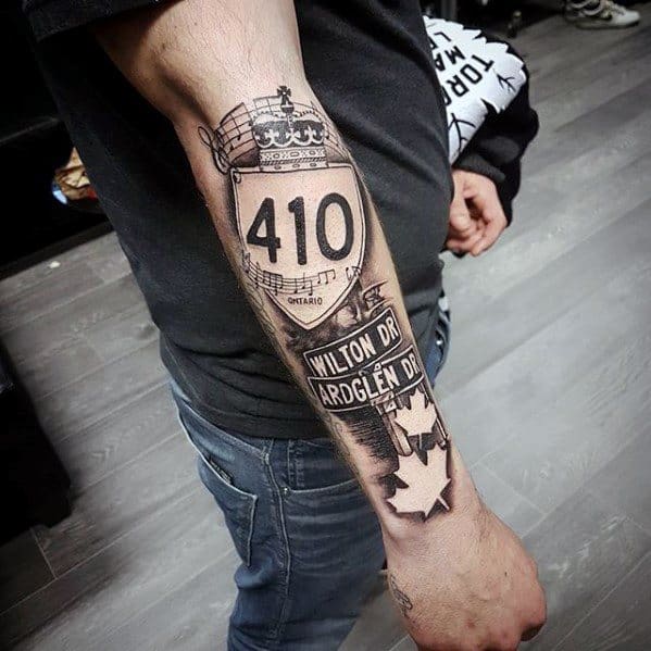 Black and grey forearm tattoo featuring Ontario Highway 410 sign, street names Wilton Dr and Ardglen Dr, music notes, and maple leaves.