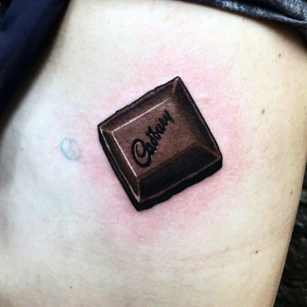 This tattoo showcases a realistic, 3D-style chocolate square with the "Cadbury" logo, giving it a lifelike and delicious appearance.