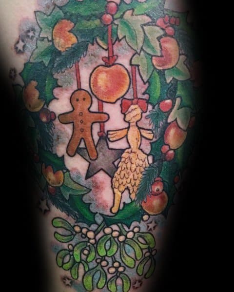 Christmas tattoo of a festive wreath decorated with holly, mistletoe, ornaments, and hanging gingerbread and straw figurines for a traditional holiday design.