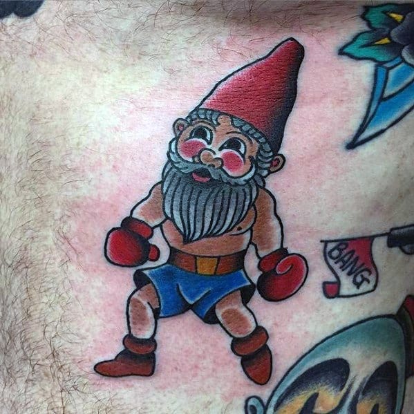 Tattoo of a muscular gnome wearing boxing gloves, blue shorts, and a red hat, in a fighting stance with a playful "BANG" flag in the background.