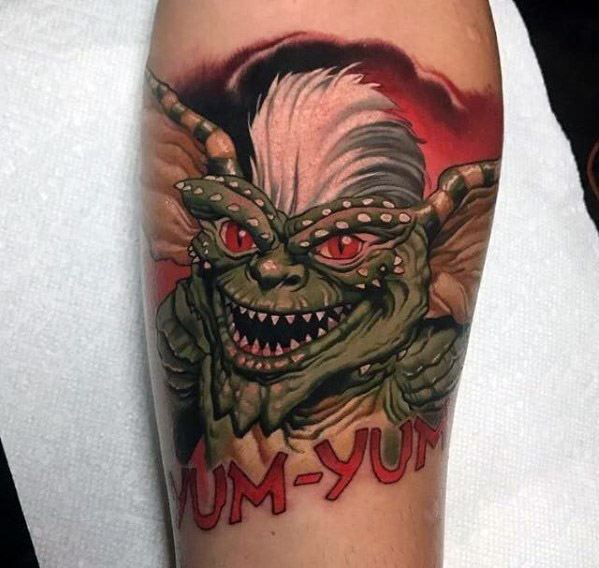 Gremlin tattoo with sharp teeth, glowing red eyes, and 'YUM-YUM' text, featuring bold colors and a fierce expression.
