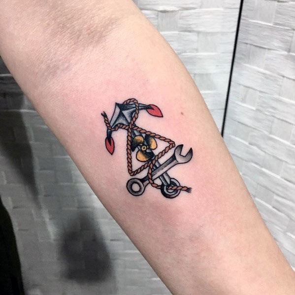 Small forearm tattoo featuring a propeller, wrench, and anchor tied together with a rope, symbolizing aviation, mechanics, and maritime themes.