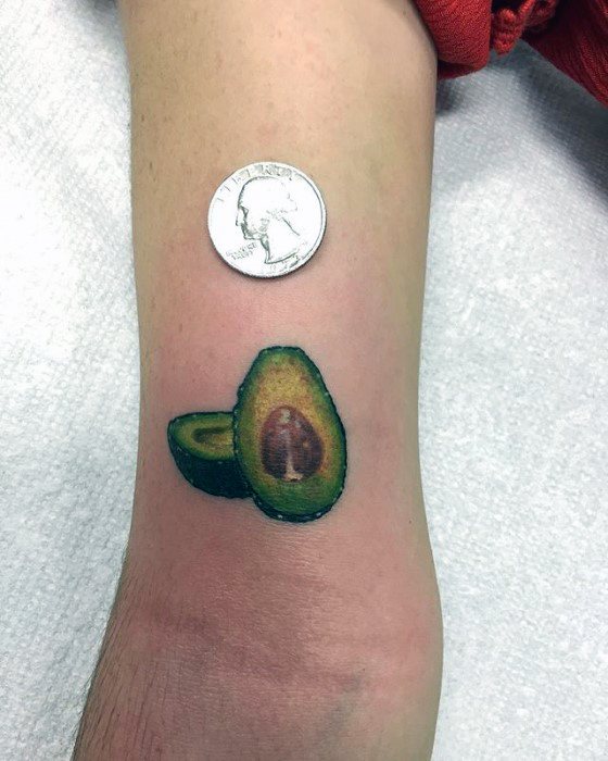 Tiny realistic avocado tattoo near the elbow, showing both halves with textured detail, placed next to a quarter for size comparison.