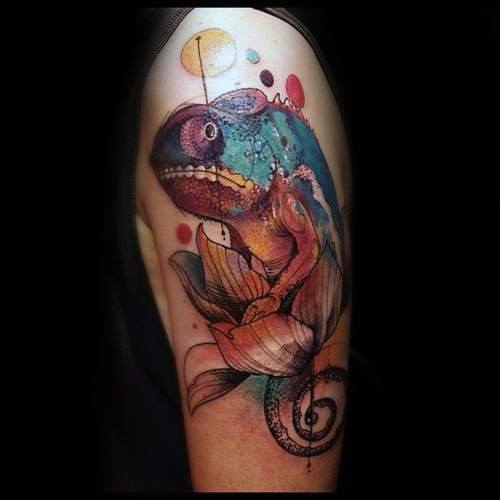 Whimsical watercolor-style chameleon tattoo with bright splashes of color, abstract dots, and a floral base on the upper arm