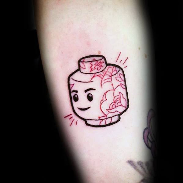 Cute tattoo of a smiling Lego head with red floral patterns and soft line details, blending playful and decorative styles on the arm.