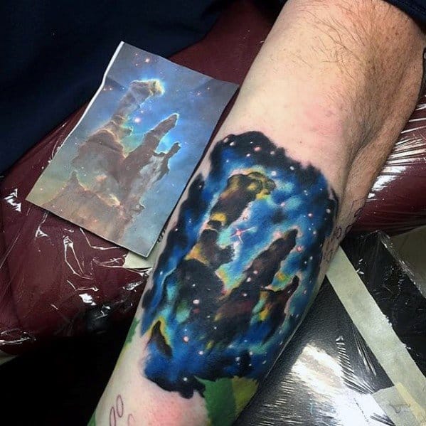 A forearm tattoo of a cosmic landscape with a nebula tattoo next to a reference image