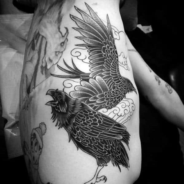 Blackwork tattoo of Huginn and Muninn in flight with stylized clouds, inked along the ribcage with bold lines and shading.
