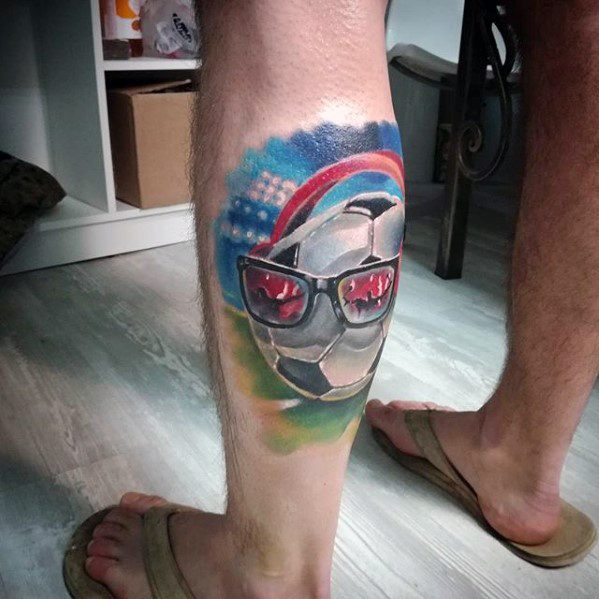 Colorful tattoo of a soccer ball wearing sunglasses with stadium lights, rainbow, and a playful vibe, inked on the back of the leg.