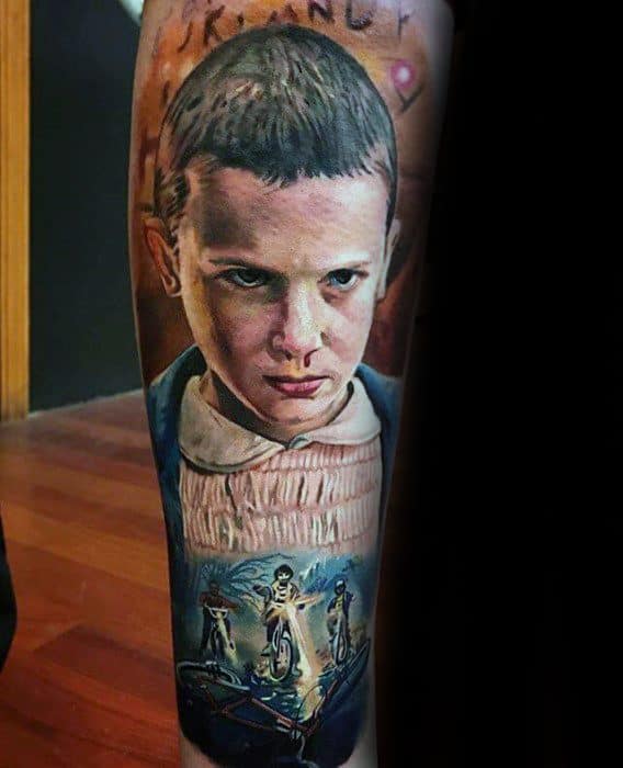 Realistic tattoo of Eleven with intense expression, featuring detailed scenes of the kids on bikes and the Demogorgon below her.