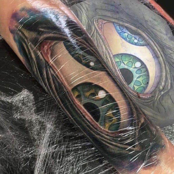 Tool-inspired forearm tattoo showing two realistic green-blue eyes peeking through a torn skin illusion with detailed texture and shading.