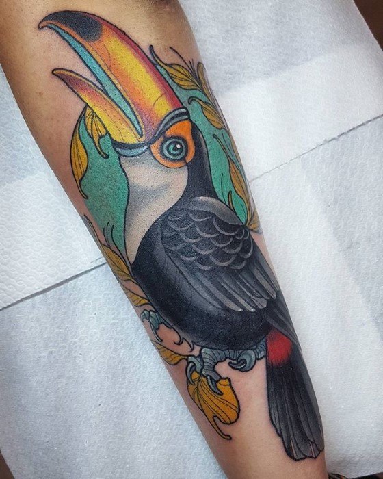 Colorful toucan tattoo on the forearm with a bold yellow-orange beak, teal background, and ornate decorative leaves.