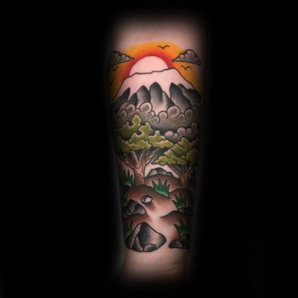 Traditional landscape tattoo with a snow-capped mountain, glowing sunset, lush trees, clouds, and a rocky path in the foreground.