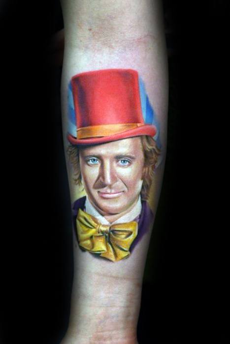 Colorful Willy Wonka tattoo with a bright red top hat, yellow bow tie, and detailed facial features set against a blue background.