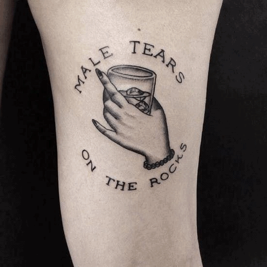 Funny tattoo of a delicate hand holding a glass with ice, surrounded by the phrase 'Male Tears on the Rocks.' A bold and witty statement.