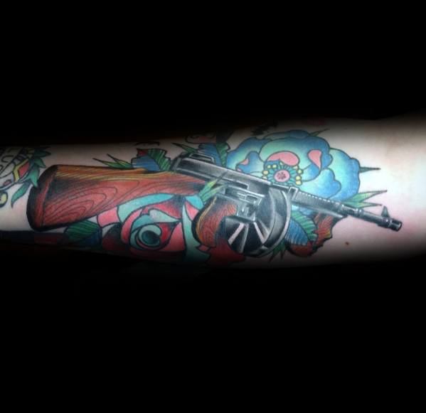 Realistic Tommy gun tattoo with detailed wood and metal textures, set against vibrant traditional-style roses for a bold contrast on the forearm.