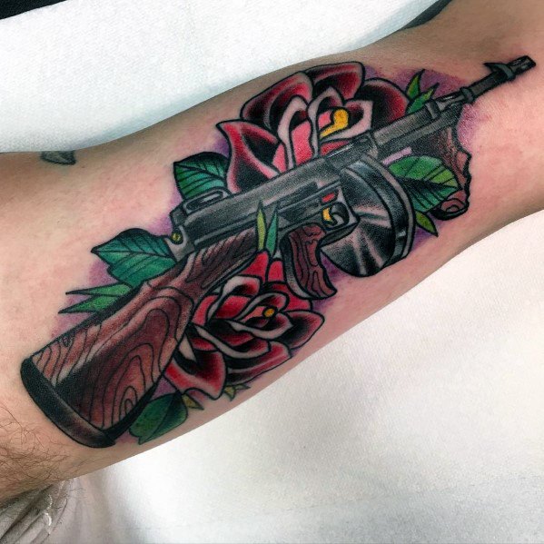 Traditional-style Tommy gun tattoo with rich wood and metal details, surrounded by bold red roses and green leaves for a striking forearm design.