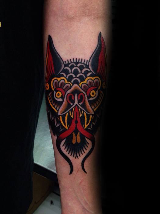 Traditional bat head tattoo on the forearm with bold black, red, and yellow ink, fierce eyes, and sharp fangs curling into tendrils.