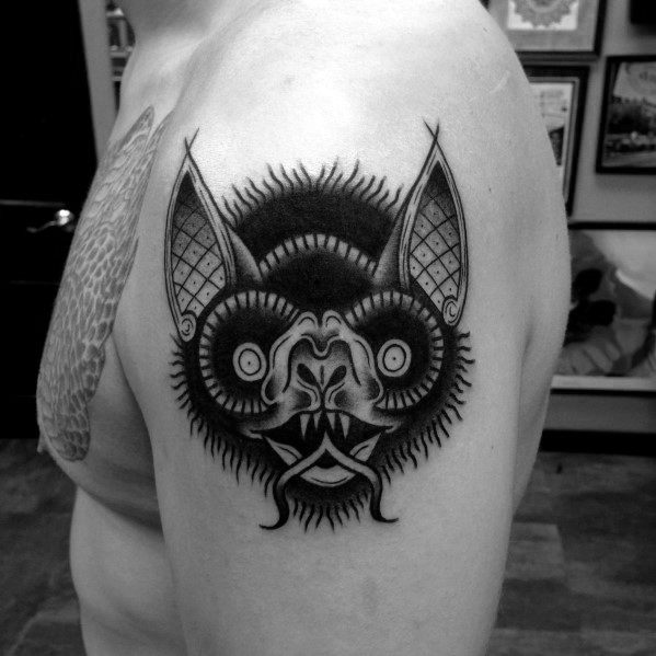 Black and grey bat head tattoo on the upper arm with bold linework, circular eyes, sharp fangs, and textured shading details.