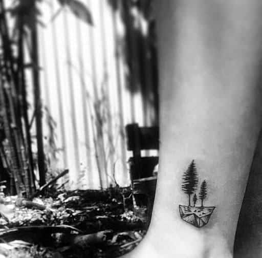 Minimalist black ink tattoo on the ankle, featuring a small floating island with pine trees, creating a simple yet artistic nature-inspired design.