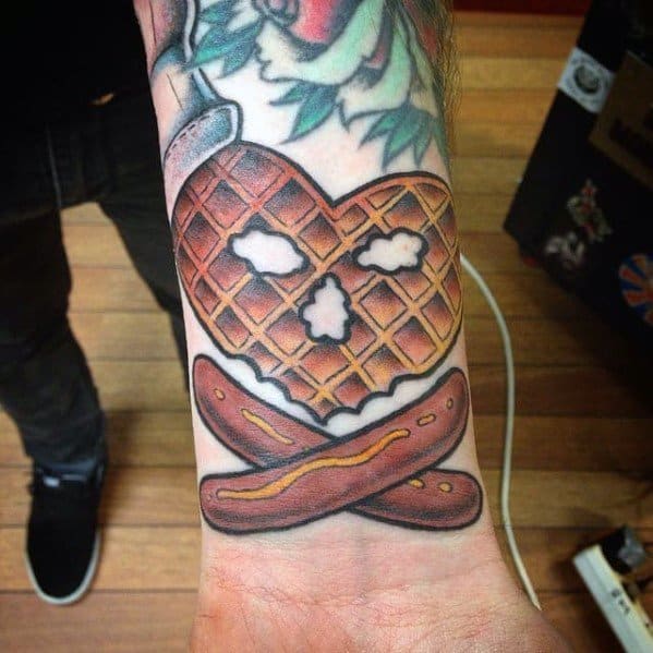 Heart-shaped waffle tattoo with a skull-like face, crossed sausages, and bold shading, blending breakfast with a rebellious and edgy design.