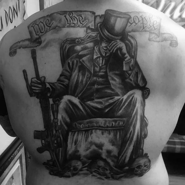Large black and grey back tattoo of a mysterious figure in a suit and top hat holding an AR-15, with a "We the People" banner above, symbolizing patriotism, power, and resistance.