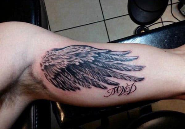 Black and grey tattoo of a detailed angel wing on the inner bicep, accompanied by elegant script initials below