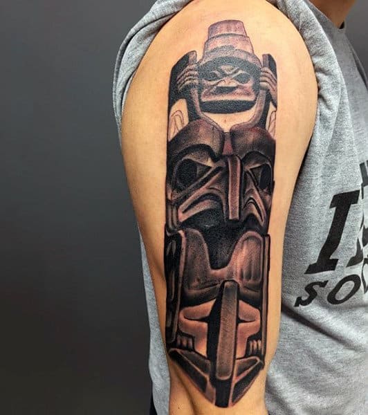 Male With Carved Dark Shaded Totem Pole On Upper Arm Tattoo