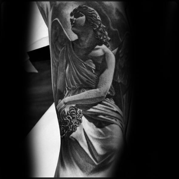 A black and grey tattoo on the forearm featuring an angel sitting with a serene expression, holding a cloud, and detailed shading on the robes and wings.