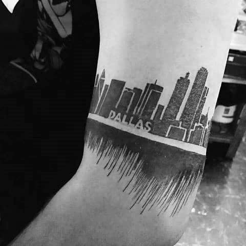 Bold Dallas skyline tattoo on the upper arm with a mirrored reflection effect and the word "DALLAS" in block letters across the center.