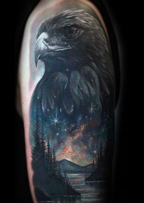 Upper arm tattoo of a detailed eagle head above a cosmic lake scene, with starry skies, pine trees, and mountains glowing in the night.