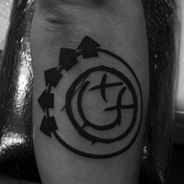 Black and white tattoo of the Blink-182 smiley face logo with bold black lines and arrows, placed on the forearm.