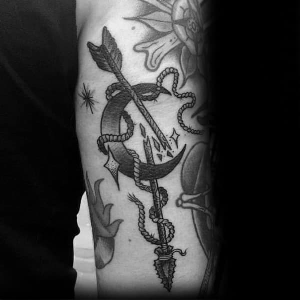 Blackwork broken arrow tattoo on the upper arm with a crescent moon, rope detail, and shards flying from the fractured center.
