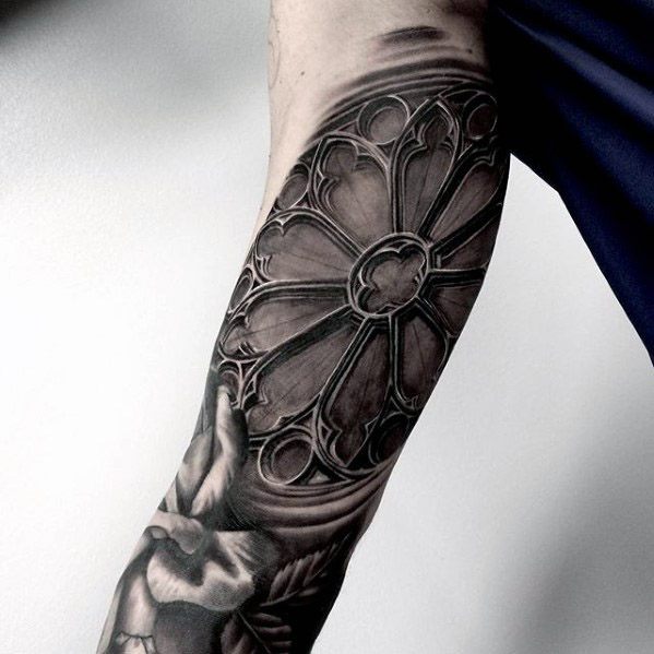 Forearm tattoo of a gothic rose window with intricate tracery and deep shading, paired with a skull and soft floral elements below.