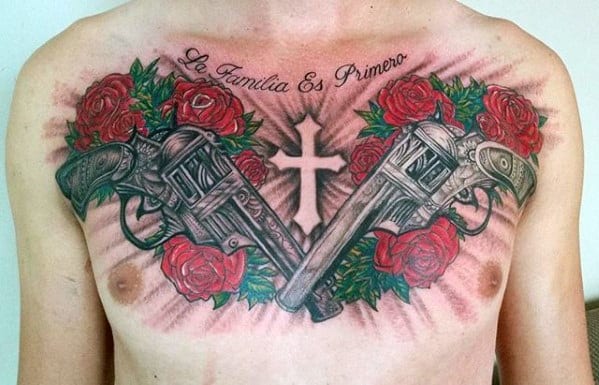 Chest tattoo with two crossed revolvers, red roses, a white cross in the center, and script reading "La Familia Es Primero" above.