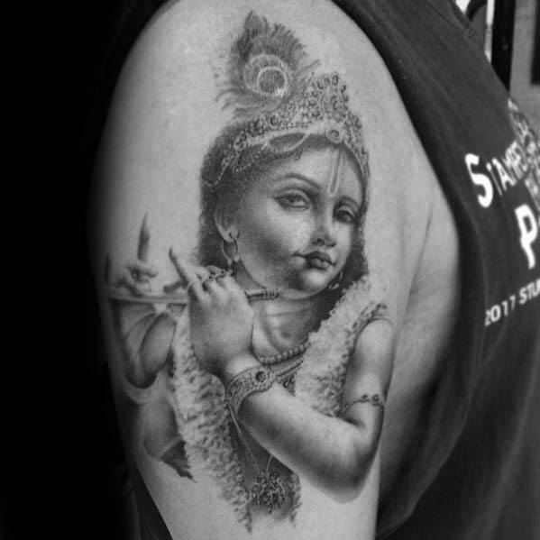 Realistic Krishna tattoo of a young Krishna playing the flute, with lifelike shading, a detailed crown, and soft facial expression on the upper arm.