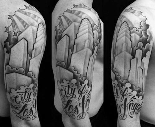 Upper arm tattoo of Los Angeles skyline with clouds, bold buildings, and cursive script reading "City of Los Angeles" in black and gray.