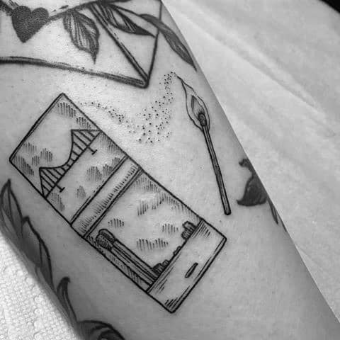 Fine line tattoo of a matchbox with a bridge and sky design, paired with a single lit match, symbolizing connection, hope, or lighting the way forward.