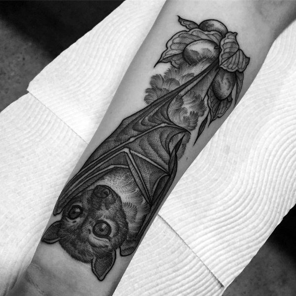Black and grey tattoo of a realistic upside-down bat with detailed shading, hanging among fruits and leaves on the forearm.