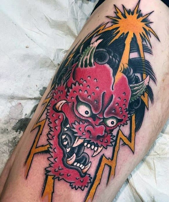 Bold Raijin thigh tattoo with intense red demon face, sharp teeth, yellow lightning bolts, and a radiant burst at the top.