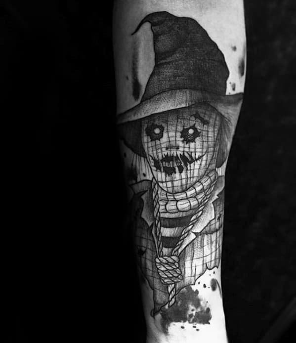 Black and grey scarecrow tattoo with button eyes, stitched smile, wide-brim hat, rope neck, and ink splatter-style background.