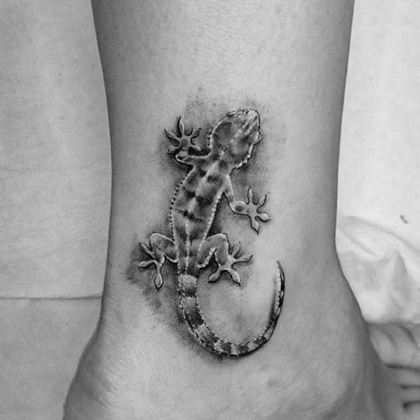 Realistic black and grey gecko tattoo on ankle with soft shading and lifelike details, appearing to crawl on the skin.