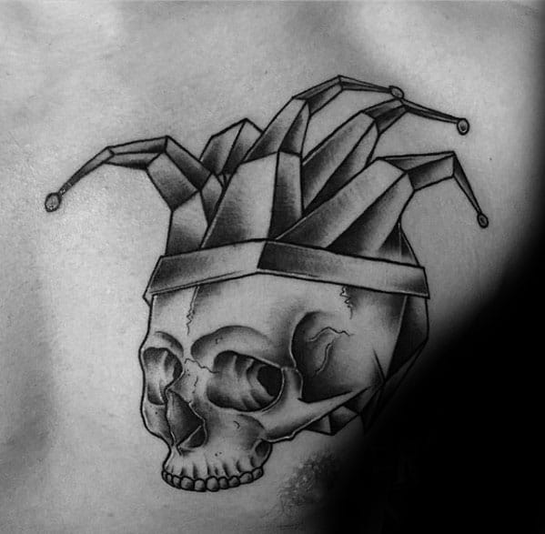 A black and gray tattoo of a skull topped with a jester hat, featuring geometric folds, placed on the upper chest.