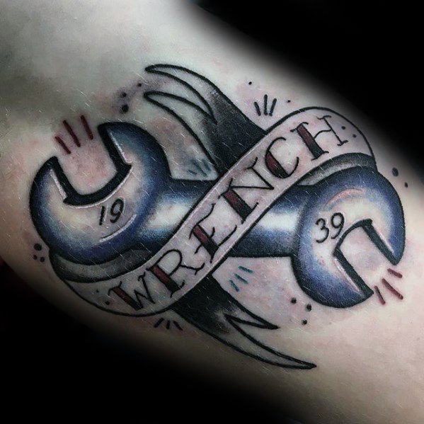 Tattoo of two blue-toned wrenches crossed with a banner that reads WRENCH and the numbers 1939, in a bold traditional style.