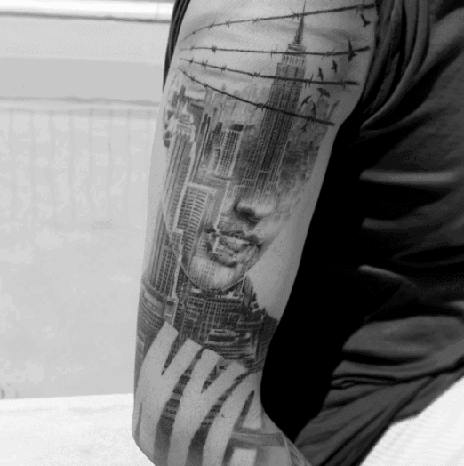 Empire State Building tattoo combined artistically with a woman's face, barbed wire, and "NYC" lettering, shaded realistically.