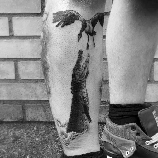 Realistic black and gray tattoo of a crocodile emerging from water, jaws open, reaching for a flying goose, inked on the lower leg.