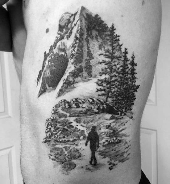 A black and gray tattoo of a lone hiker on a rugged mountain trail, surrounded by trees and towering peaks, symbolizing adventure and resilience.
