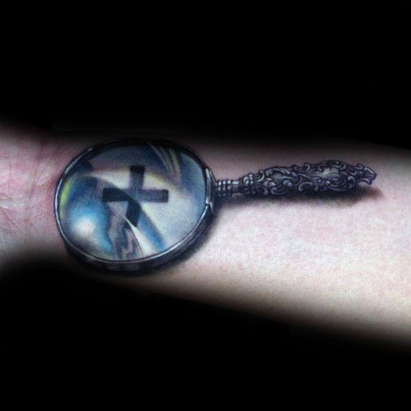 Tattoo of an ornate magnifying glass reflecting a cross and a sky scene, designed with realistic shading and color for a symbolic and spiritual look.