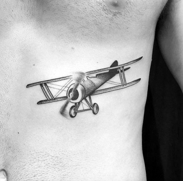 Black and grey ribcage tattoo of a vintage biplane in flight, with fine linework, shading, and a motion effect on the spinning propeller.